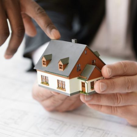 Close up shot of Caucasian contractor holding real estate project while his African colleague pointing finger at scale model building, explaining design during presentation meeting in office