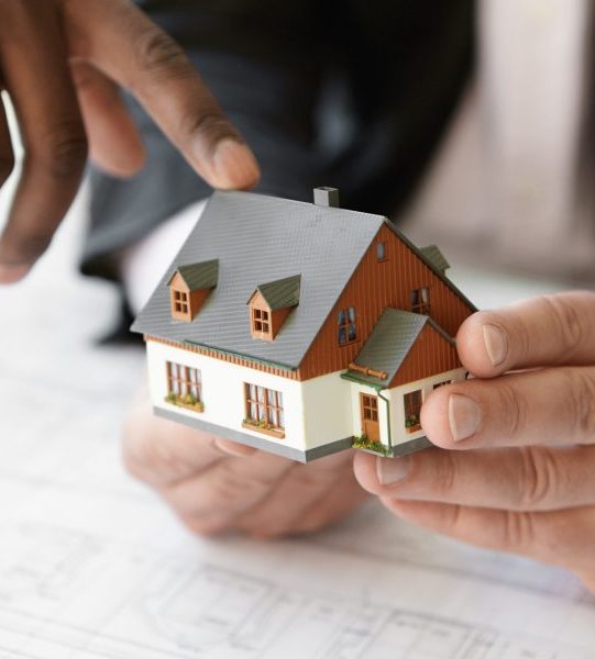 Close up shot of Caucasian contractor holding real estate project while his African colleague pointing finger at scale model building, explaining design during presentation meeting in office