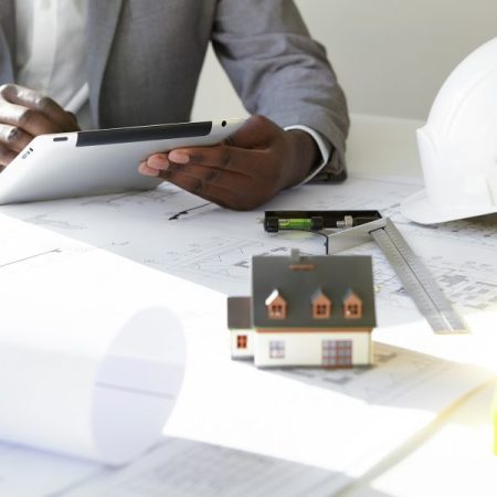 Cropped shot of dark-skinned contractor holding touch pad, entering data while working on new housing project, sitting at desk with drawings, scale model house, blueprint rolls, ruler and helmets